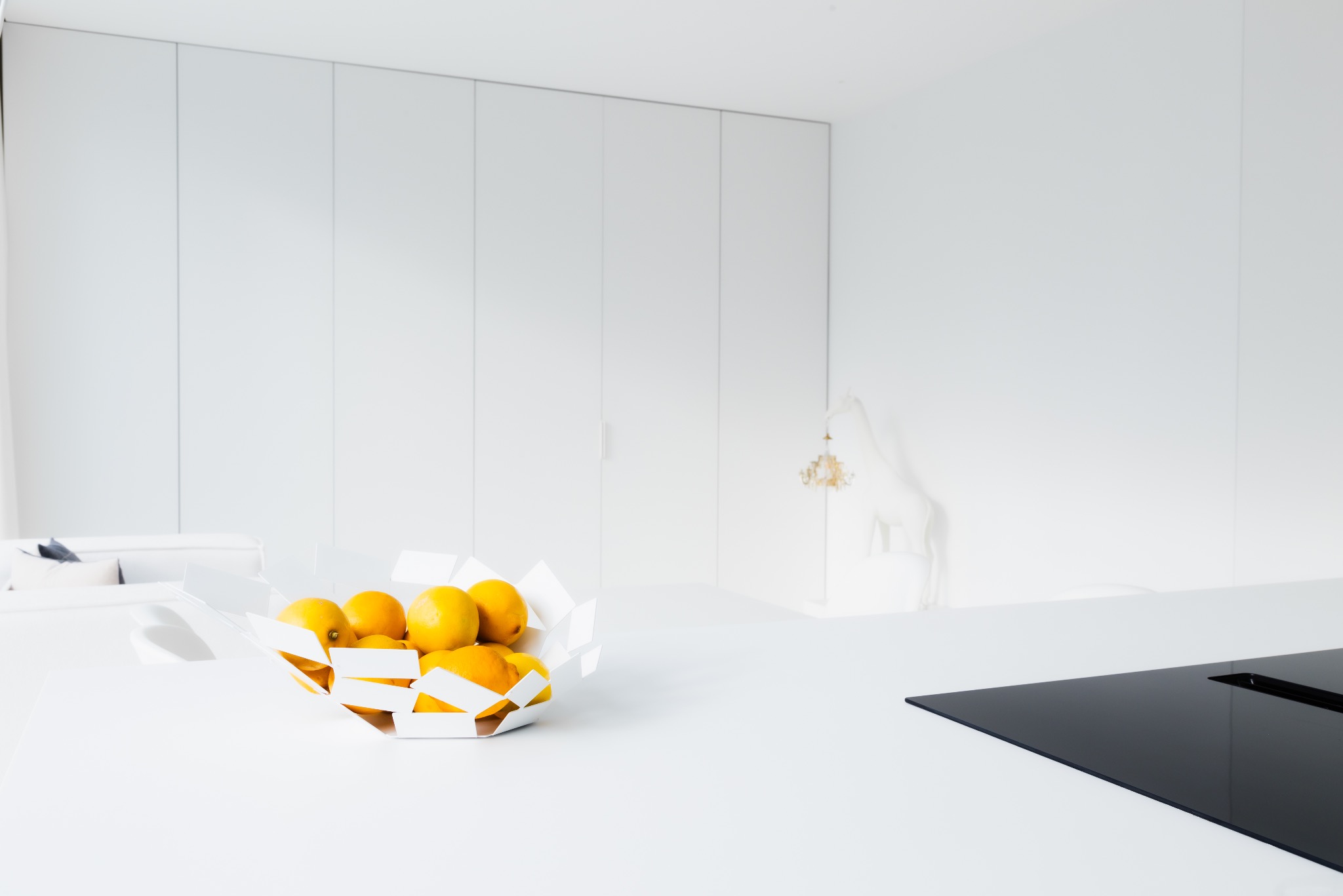 Minimalistyczna, biała kuchnia z owocami w designerskiej misie na blacie, widok na zabudowę meblową i dekoracyjną figurę konia.