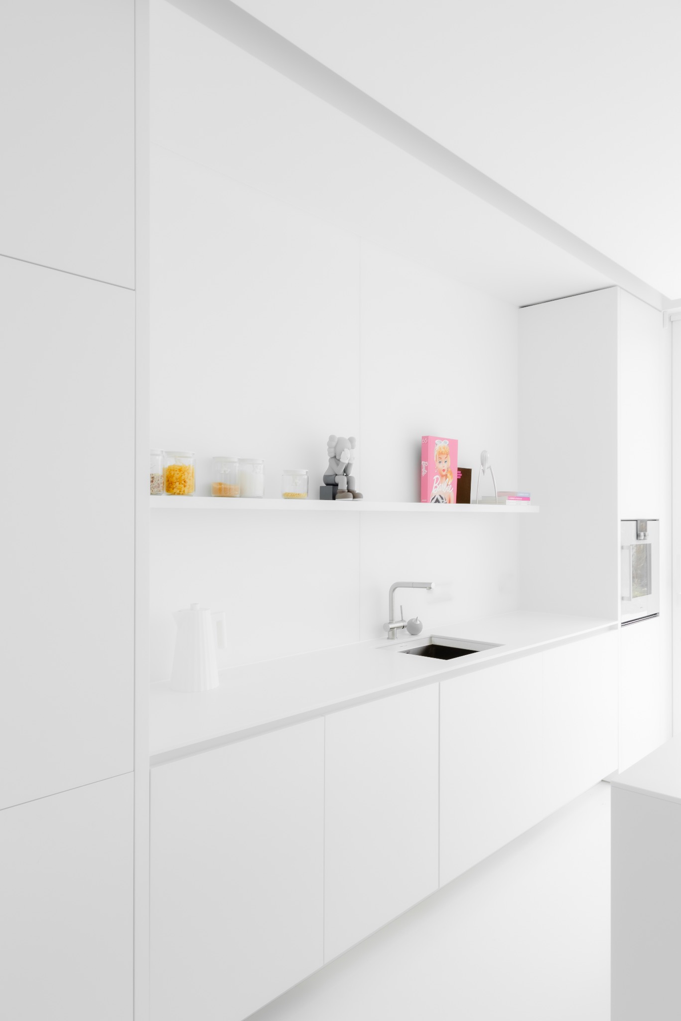 Minimalistyczna, całkowicie biała kuchnia z blatem, szafkami, półką z dekoracjami, zlewozmywakiem i piekarnikiem w zabudowie.