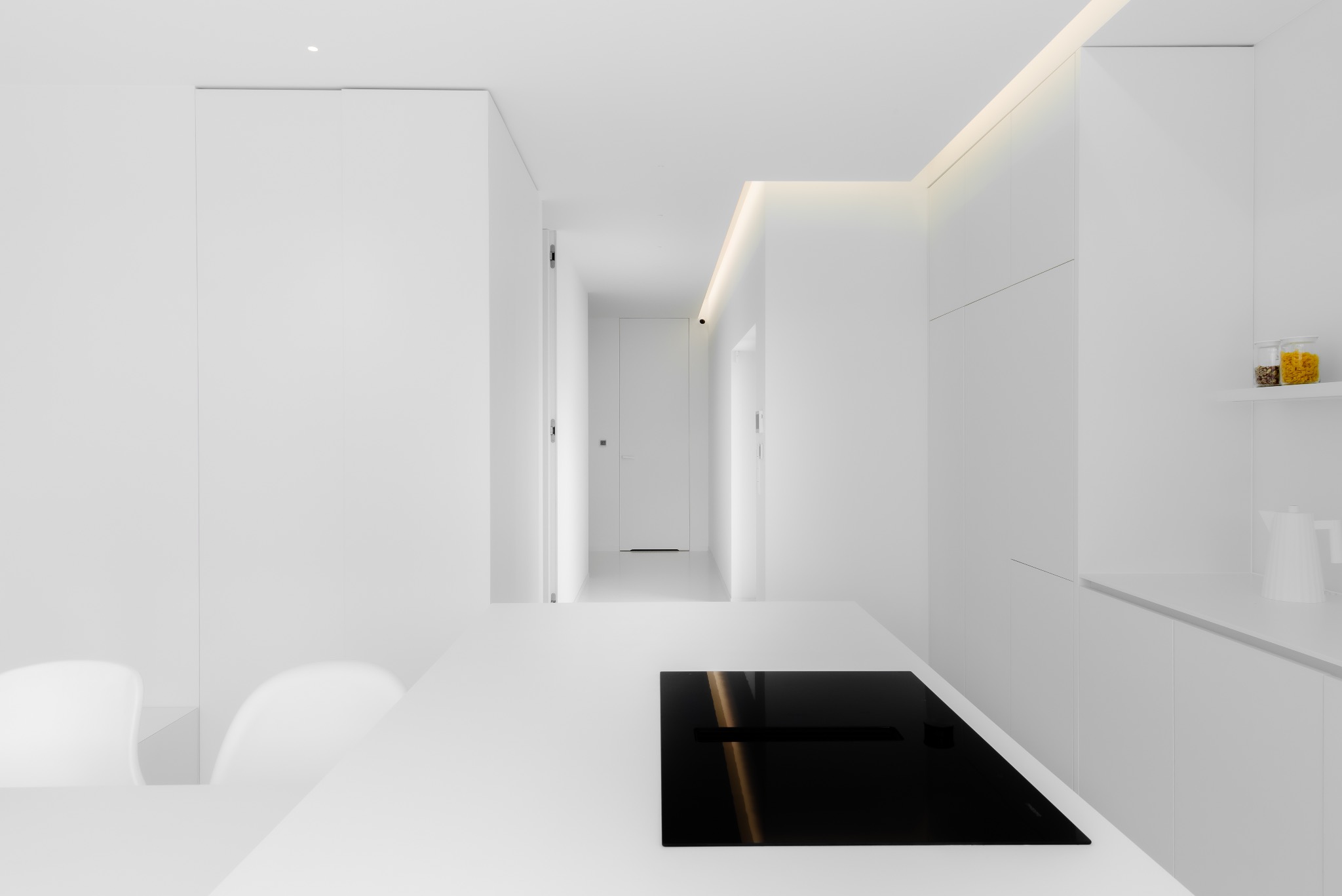 Minimalistyczna, biała kuchnia z czarną płytą indukcyjną na wyspie, widok na korytarz z białymi drzwiami i oświetleniem LED.