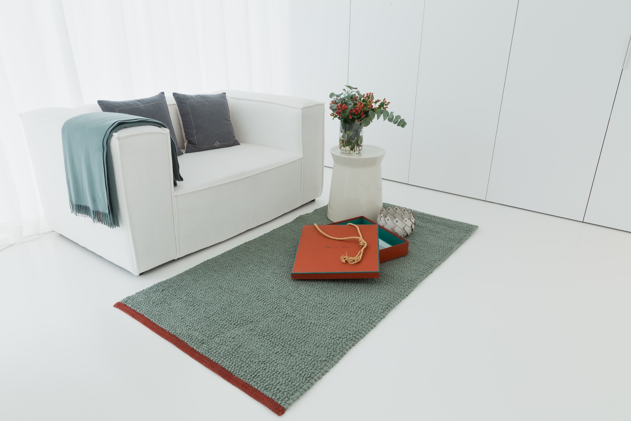 Minimalistyczne wnętrze z białym fotelem, szarymi poduszkami, zielonym pledem, dywanem z czerwonym obramowaniem, stolikiem z bukietem i pomarańczowym pudełkiem z zawartością.