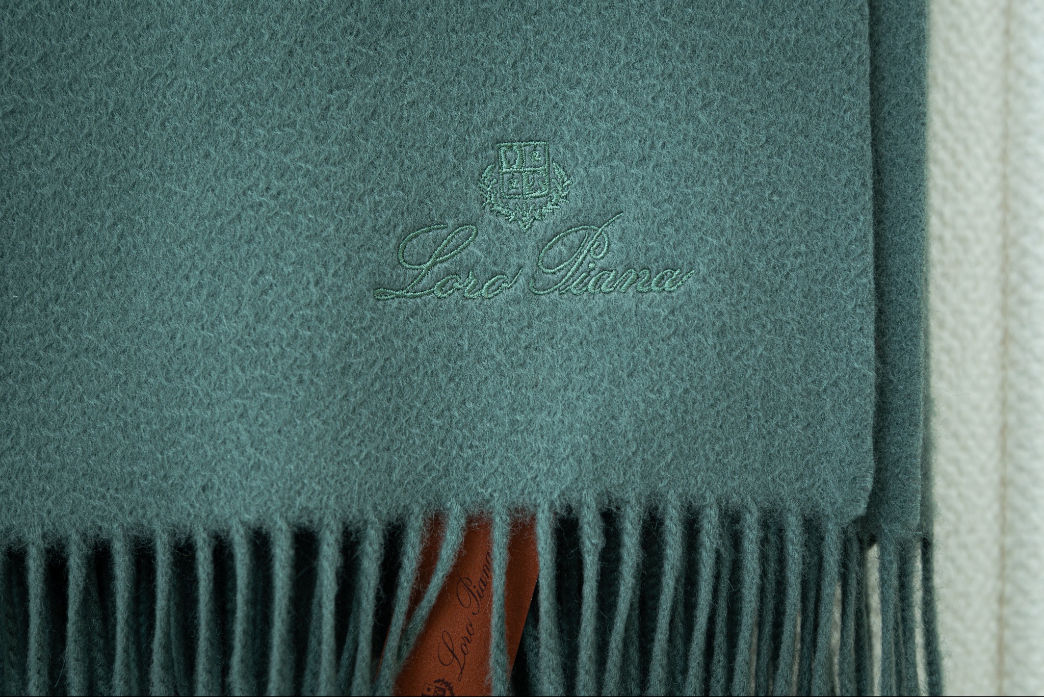 Turkusowy szal z kaszmiru z delikatnym haftowanym logo Loro Piana i frędzlami, leżący na kremowym tle.