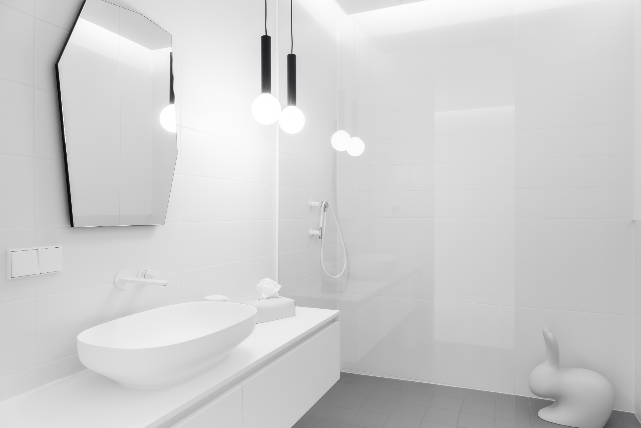 Minimalistyczna, biała łazienka z owalną umywalką, geometrycznym lustrem, wiszącymi lampami i dekoracyjną figurką królika.