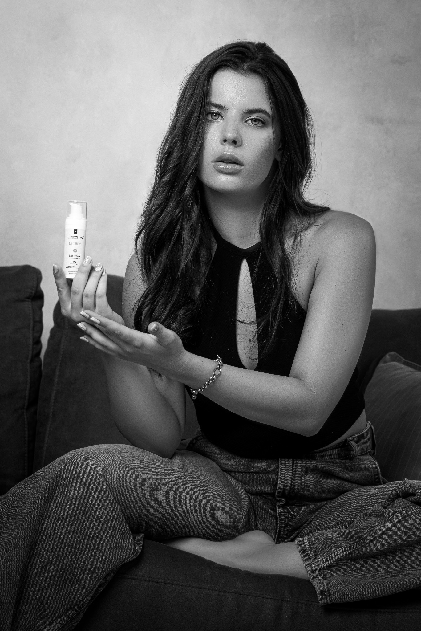 Czarno-białe zdjęcie kobiety siedzącej na kanapie, prezentującej białą buteleczkę kosmetyku do pielęgnacji twarzy, z widocznym napisem 'Lift Yeux' i dłońmi z jasnym manicure.