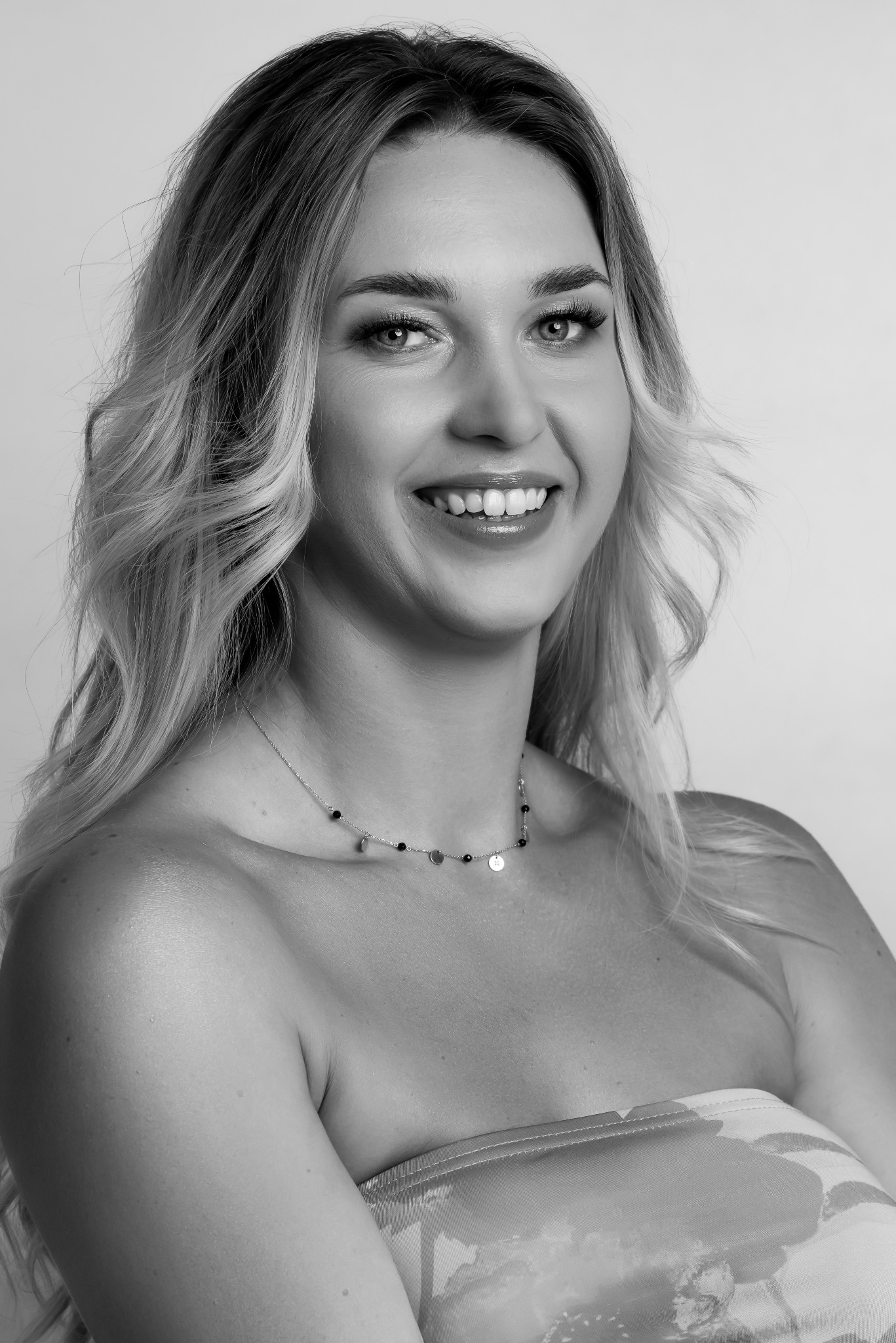 Portret uśmiechniętej kobiety w Warszawie, w czarno-białej tonacji, z delikatnym makijażem i falującymi blond włosami, noszącej subtelny naszyjnik.
