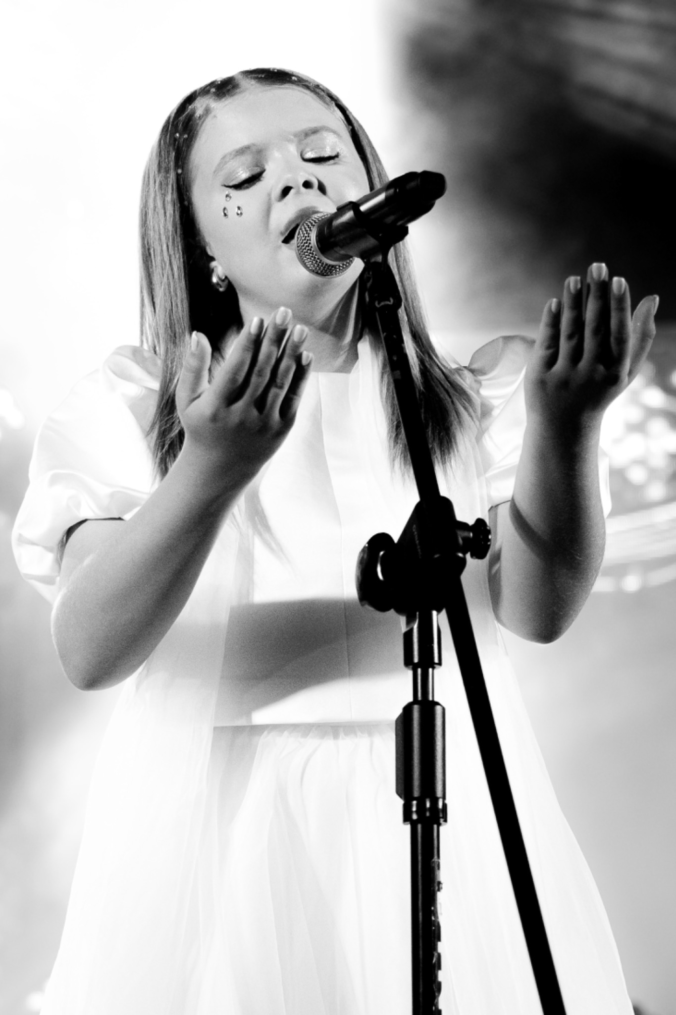 Czarno-białe zdjęcie młodej wokalistki śpiewającej do mikrofonu na statywie, z dłońmi uniesionymi w geście ekspresji, z błyszczącymi ozdobami na twarzy i we włosach, na rozmytym tle sceny.