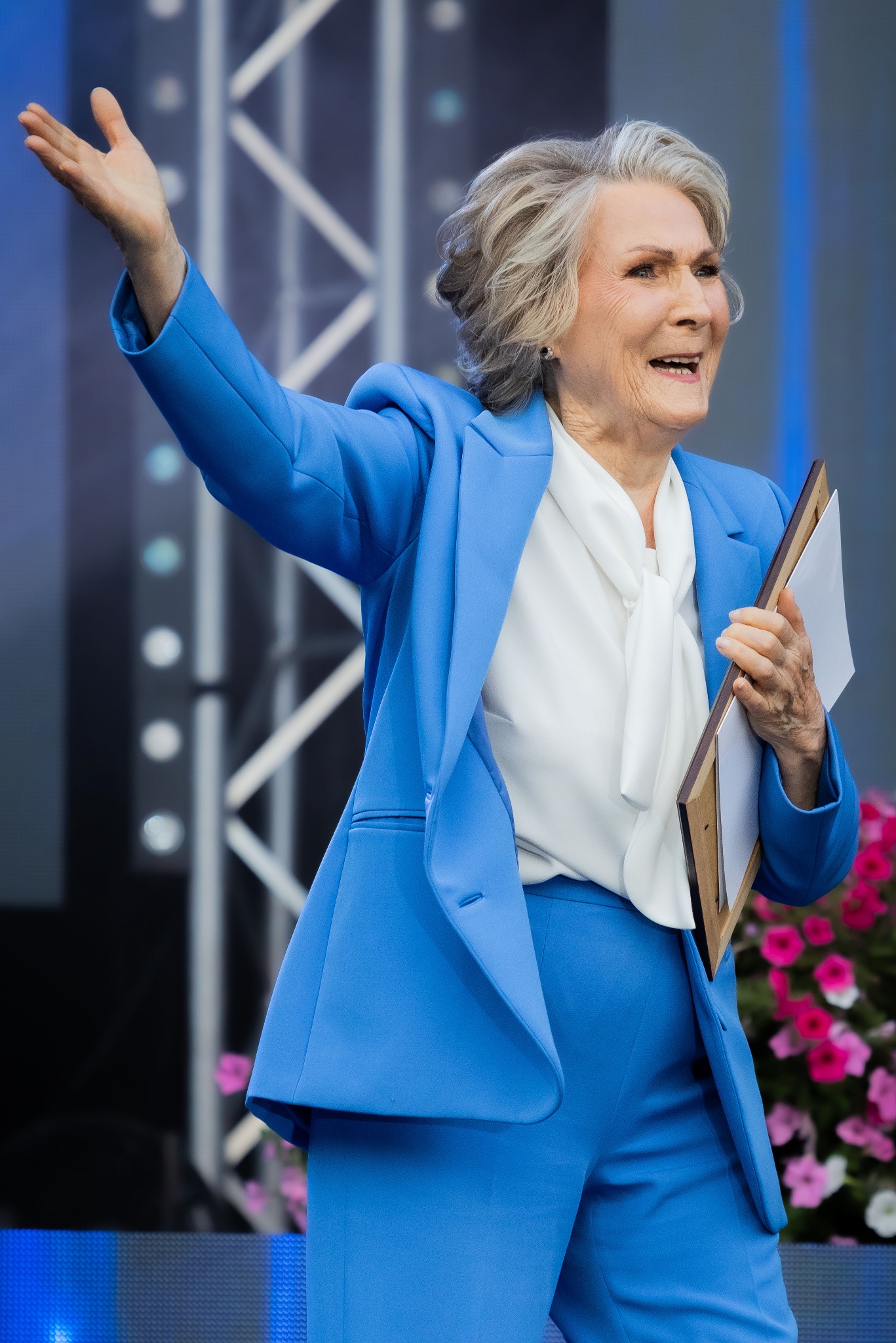 Uśmiechnięta kobieta w niebieskim garniturze, trzymająca oprawiony dokument i gestykulująca ręką na tle dekoracji kwiatowych.