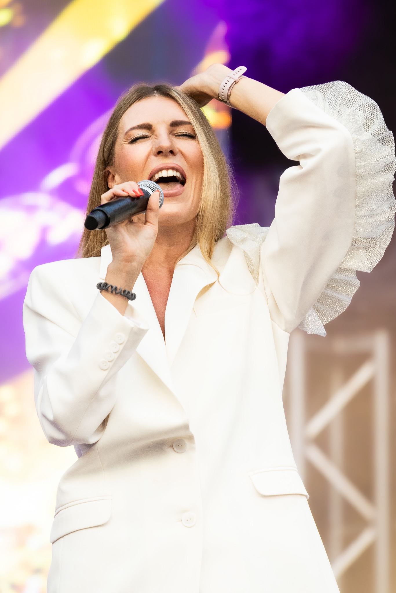 Piosenkarka w białym garniturze śpiewa do mikrofonu na scenie z fioletowym oświetleniem, unosząc jedną rękę do góry.