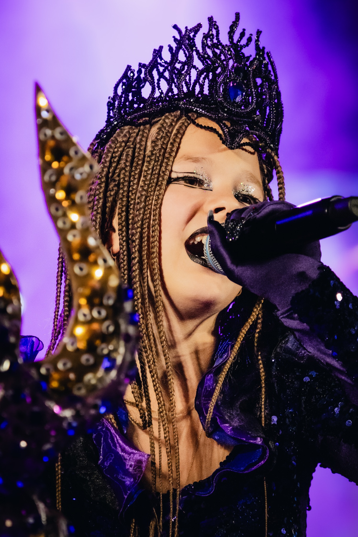 Młoda wokalistka z warkoczykami, w czarnej koronie i cekinowej sukience, śpiewa do mikrofonu z brokatem na powiekach na fioletowym tle.