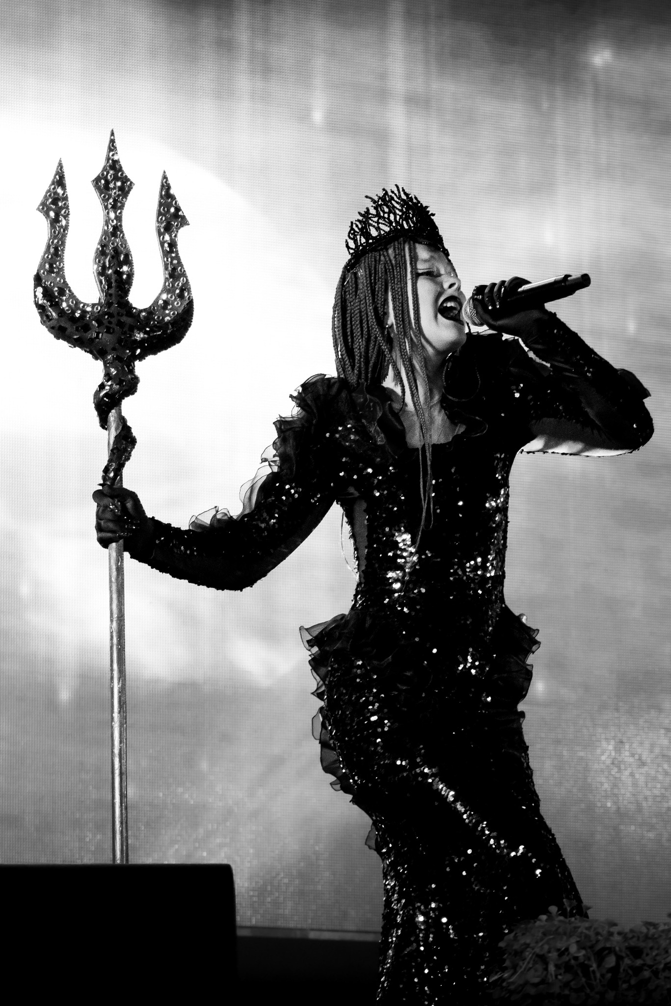 Czarno-białe zdjęcie kobiety w cekinowej sukni i koronie, trzymającej trójząb i śpiewającej do mikrofonu na scenie z rozmytym tłem.