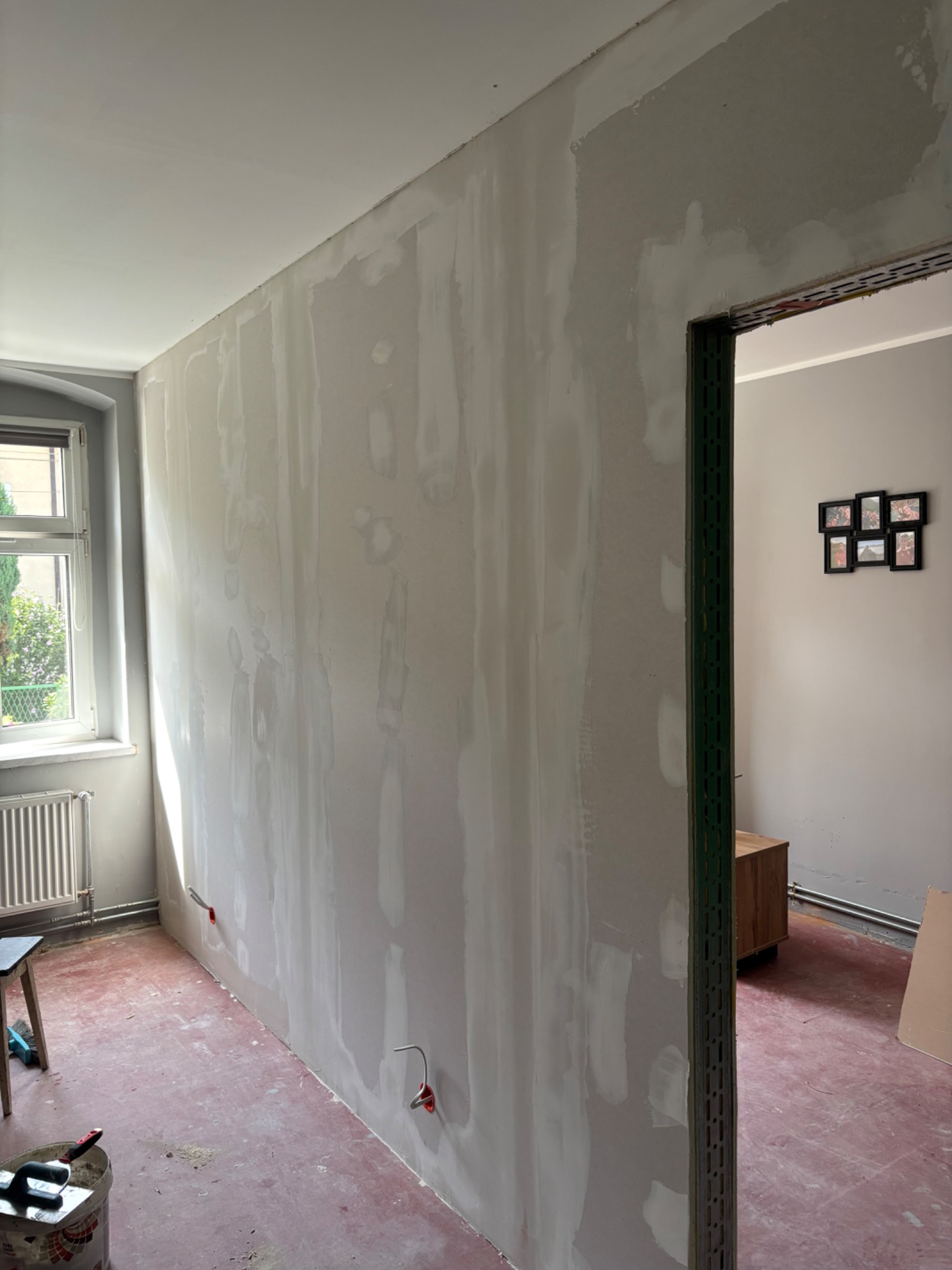 Wnętrze w trakcie szpachlowania: ściana z widocznymi warstwami gładzi, okno z grzejnikiem, narzędzia malarskie i przygotowane kable elektryczne w ścianie.