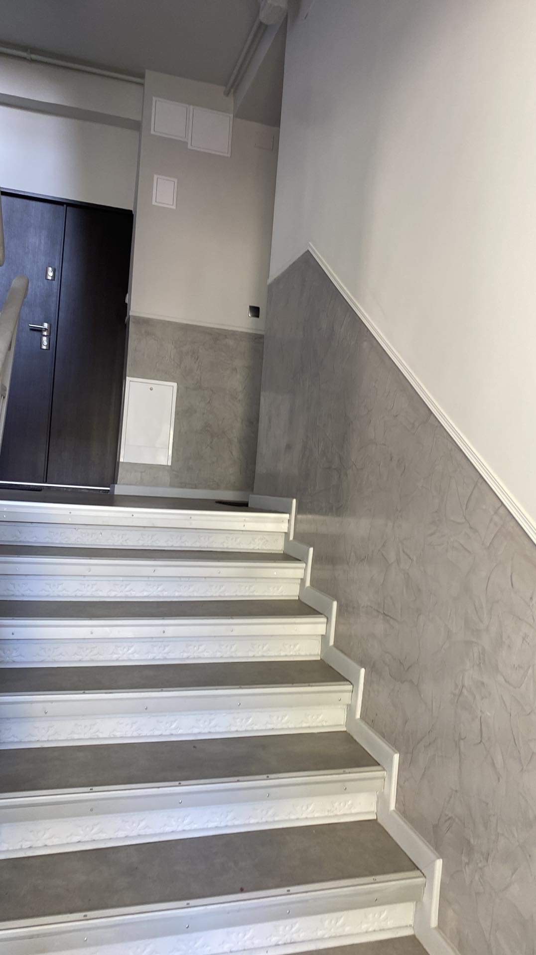 Klatka schodowa po remoncie: białe stopnie z szarymi powierzchniami, ściany z szarą fakturą do połowy wysokości i białe powyżej, ciemne drzwi wejściowe.
