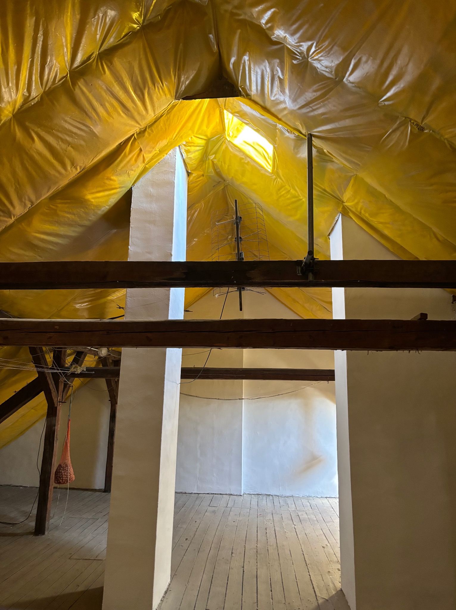 Poddasze z drewnianą konstrukcją, ocieplone żółtą folią, z widoczną anteną i fragmentem okna dachowego. Ściany i podłoga w jasnych barwach.