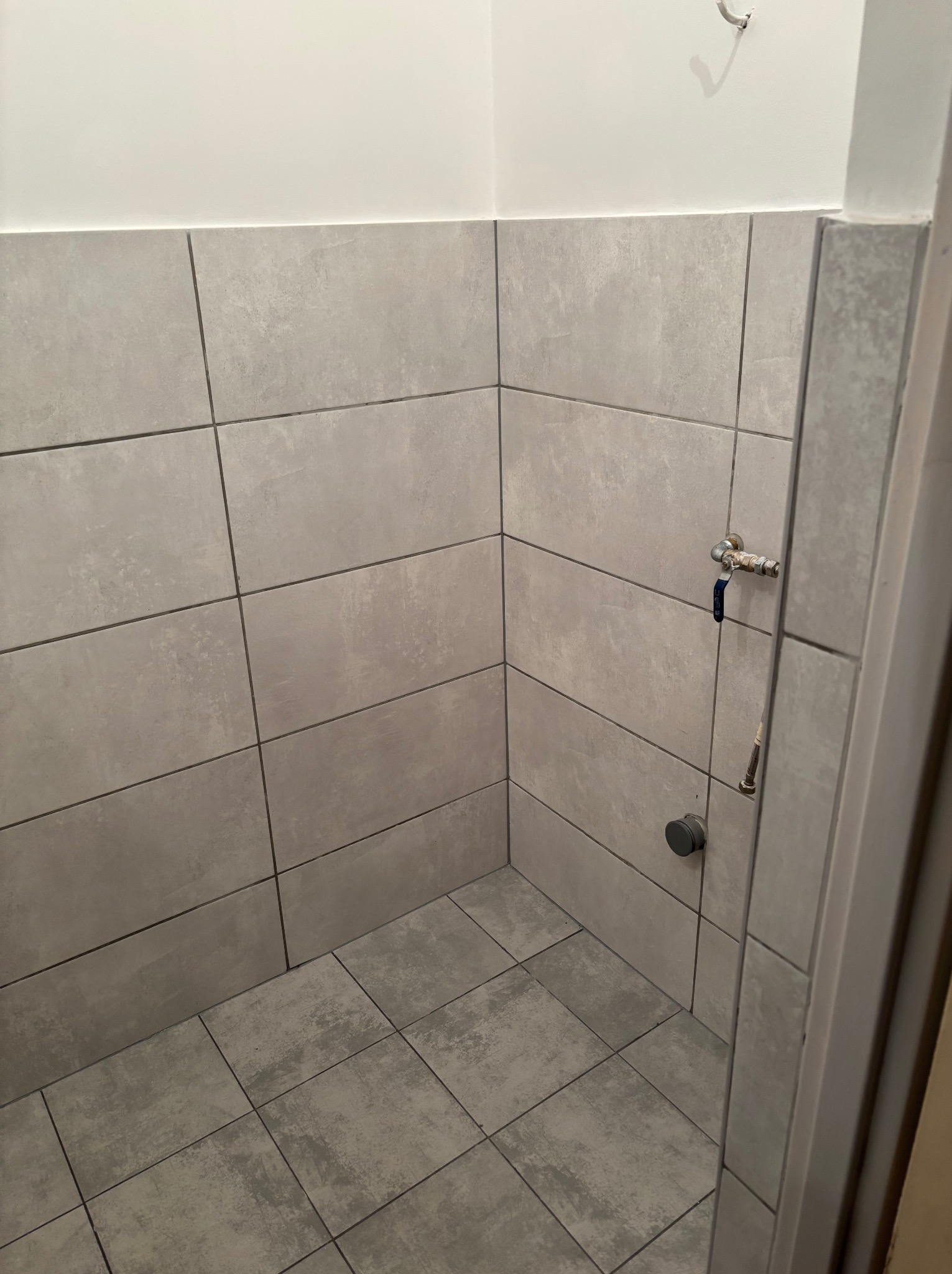 Wykończona łazienka z szarymi, matowymi płytkami na ścianach i podłodze. Widoczny zawór wody i haczyk na ścianie, minimalistyczny design.