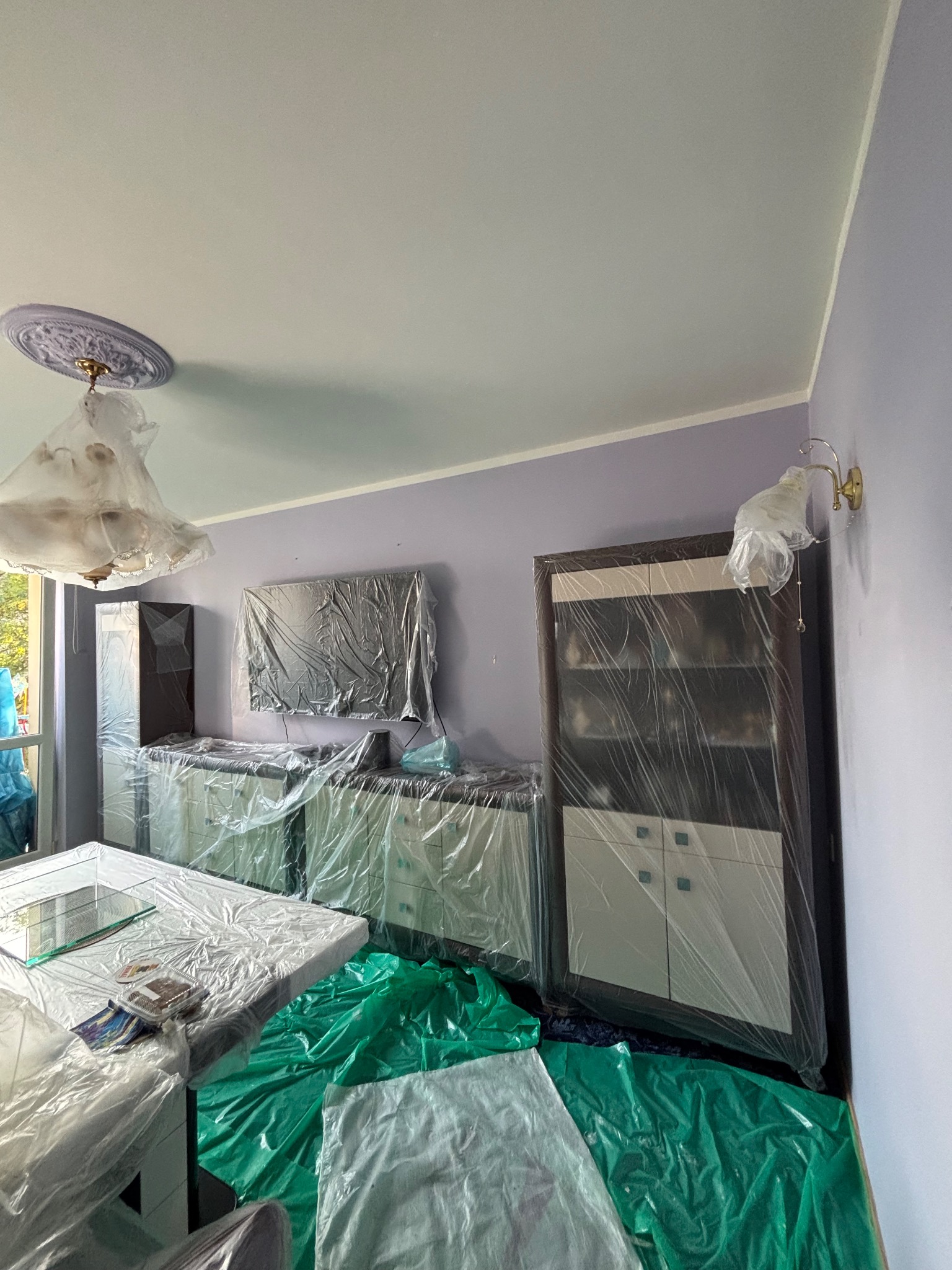 Pokój z meblami zabezpieczonymi folią malarską, ściany w odcieniach fioletu, przygotowany do prac wykończeniowych. Widoczny żyrandol i lampa ścienna.