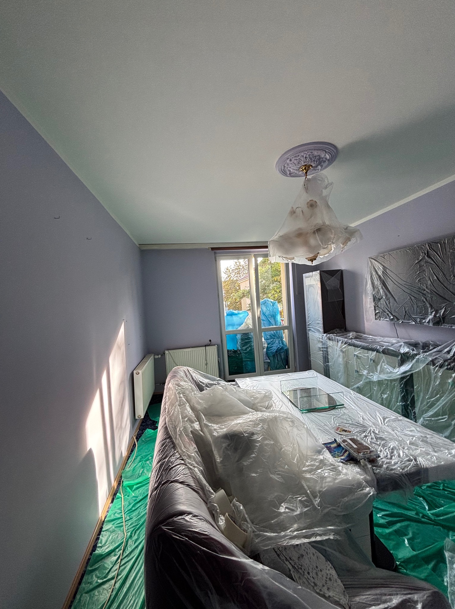 Pokój przed malowaniem: meble i podłoga zabezpieczone folią, ściany w odcieniach fioletu, widoczne promienie słoneczne wpadające przez okno. Lampa sufitowa zakryta folią.