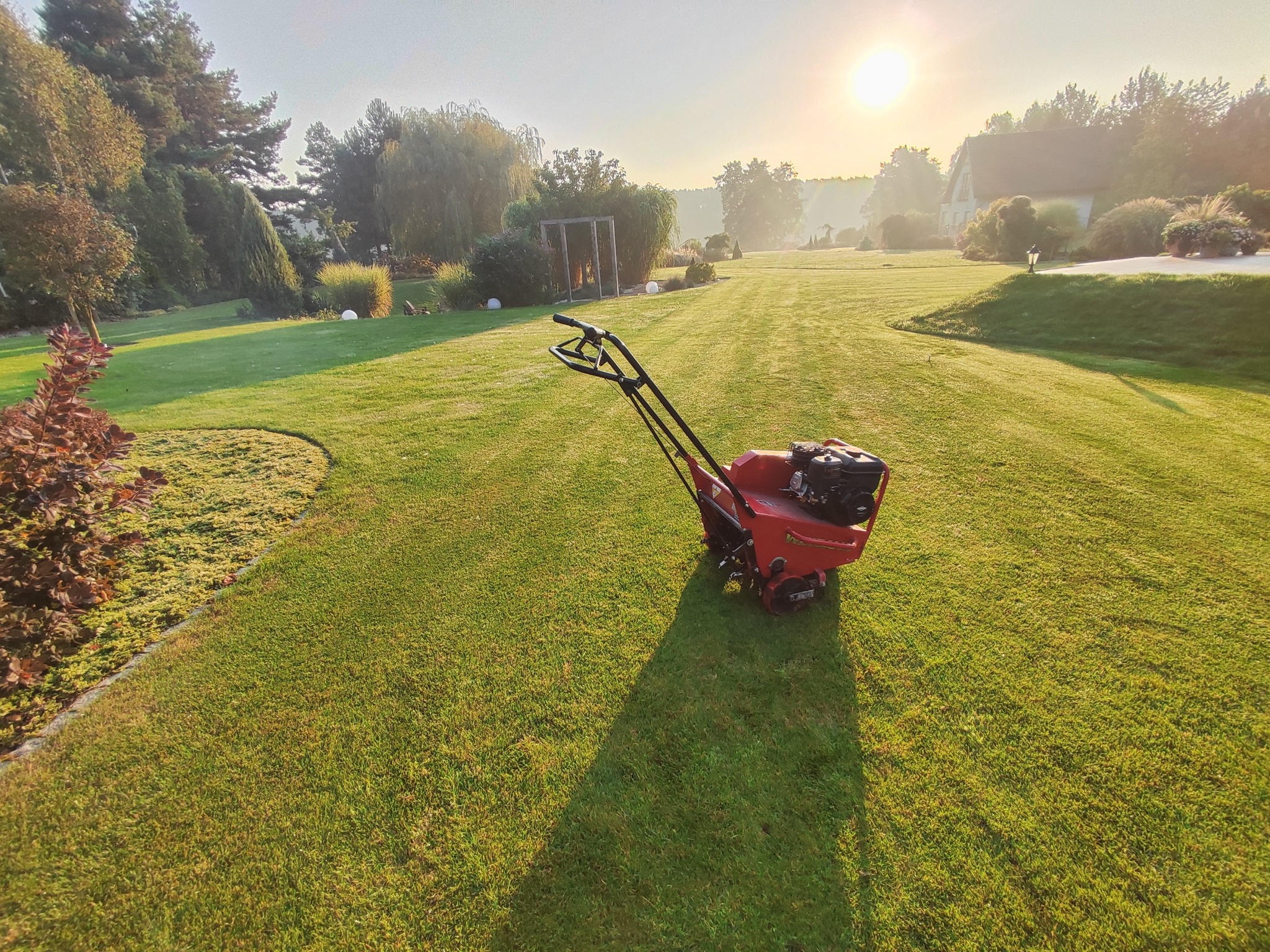 Czerwony aerator spalinowy na zadbanym trawniku w słoneczny poranek, widok na rozległy ogród z drzewami i budynkiem w tle.