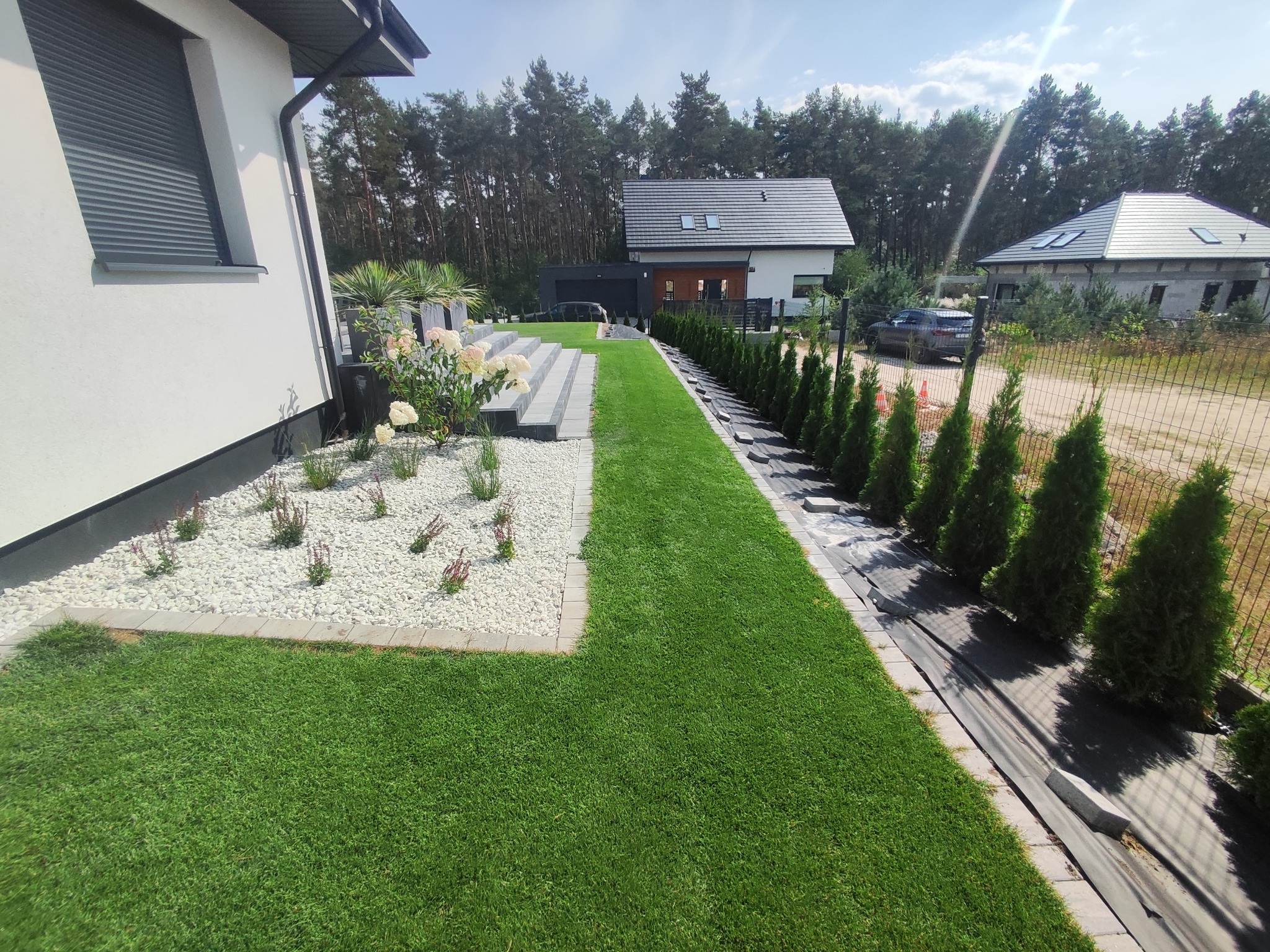 Nowoczesny ogród z równo przystrzyżonym trawnikiem, białymi kamieniami i roślinami ozdobnymi wzdłuż domu, schody z szarych płyt prowadzące do wyżej położonej części ogrodu, rząd młodych tui wzdłuż...