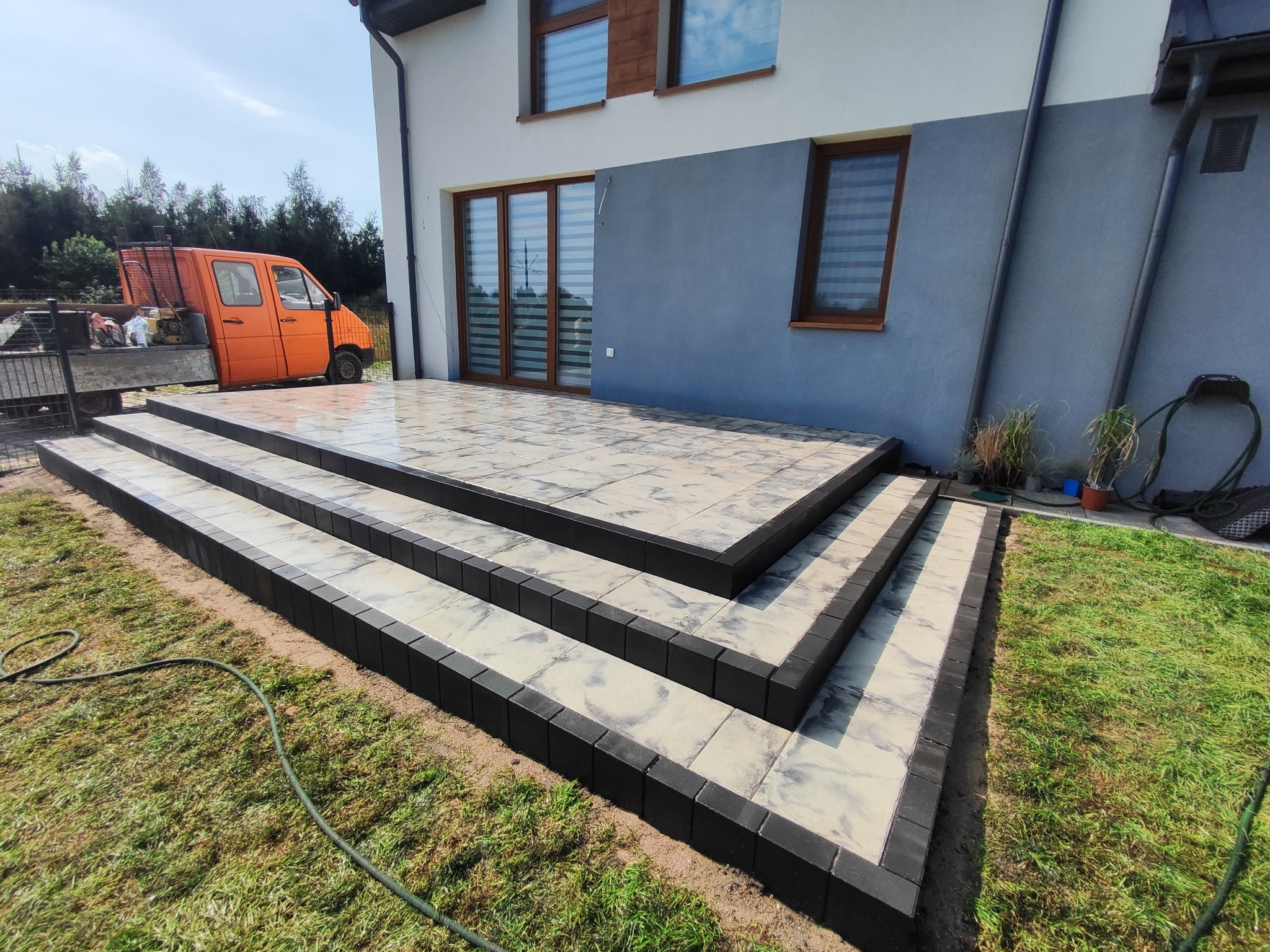 Nowoczesne schody tarasowe z kostki brukowej w odcieniach szarości i czerni, prowadzące do domu z szarą elewacją i pomarańczowym samochodem dostawczym w tle.