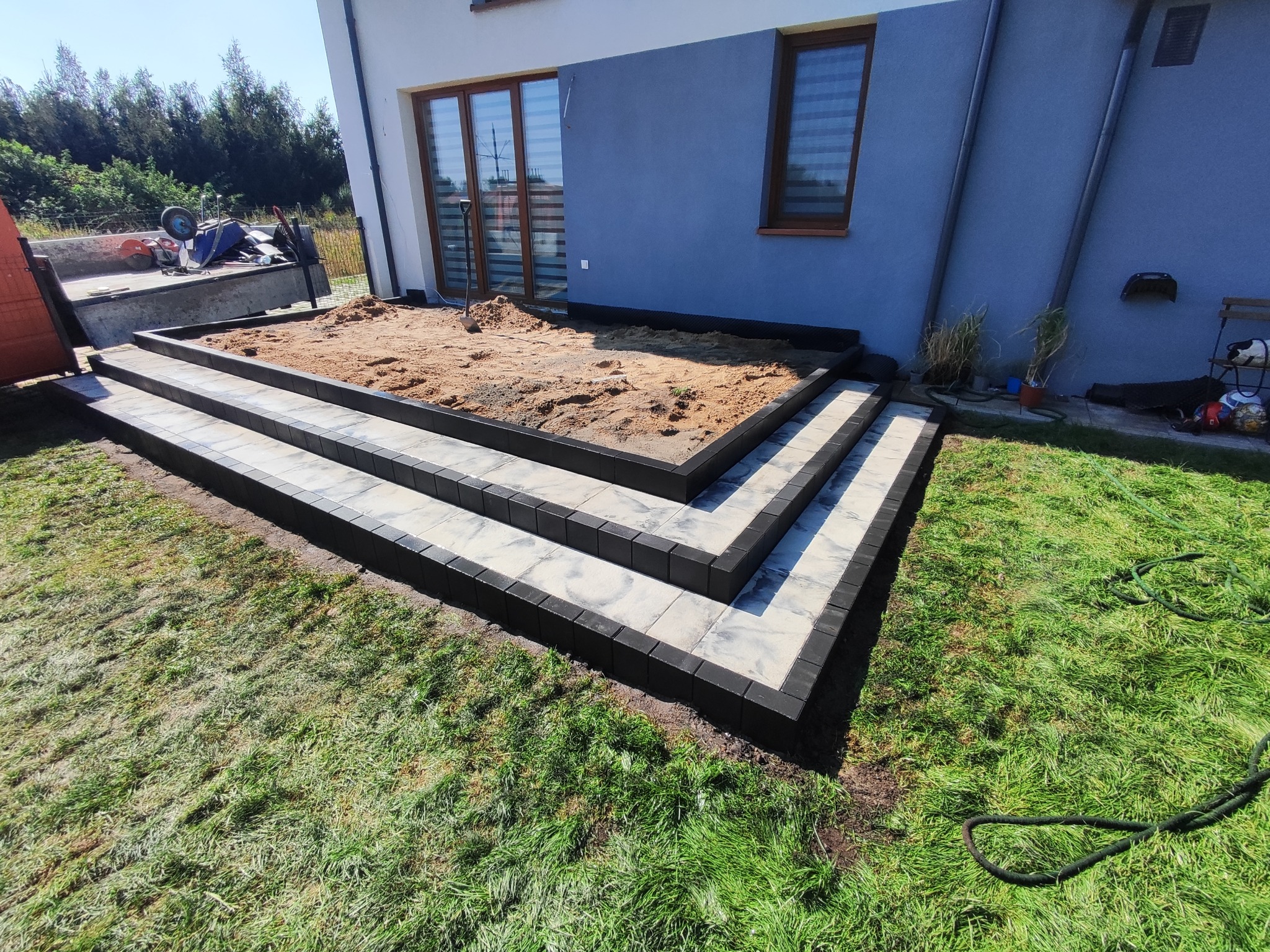 Budowa schodów z kostki brukowej prowadzących do tarasu z piaskiem, trawa w tle, widoczne narzędzia i materiały budowlane w przyczepie.