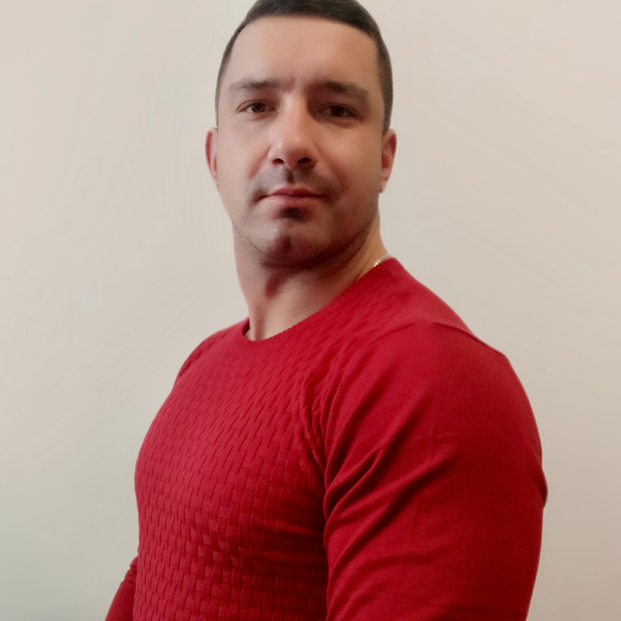 Mężczyzna w czerwonym swetrze, z krótkimi ciemnymi włosami, patrzy w obiektyw. Neutralne tło, oświetlenie dzienne. Widoczny fragment srebrnego łańcuszka.