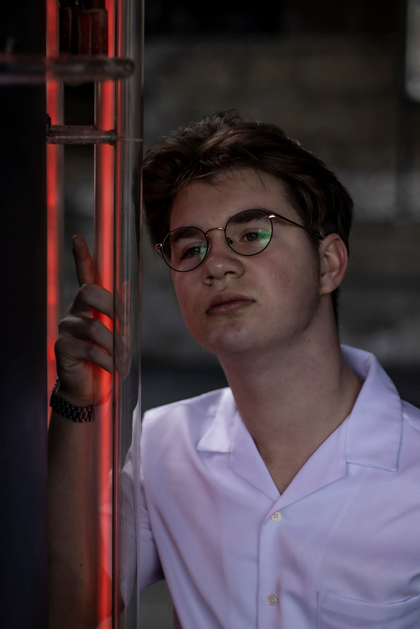 Młody mężczyzna w okularach i białej koszuli pozuje opierając się o metalową konstrukcję z czerwonym neonowym oświetleniem.