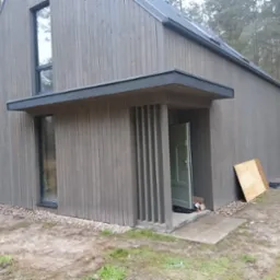 Nowoczesny dom szkieletowy z elewacją z desek w odcieniach szarości, widoczne pionowe elementy dekoracyjne przy wejściu i fragment dachu z blachy na tle lasu.