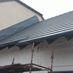 Usługi Ciesielsko-Dekarskie Daria Nawrocka - Nowy dach z ciemnej dachówki ceramicznej, widoczne obróbki blacharskie na styku z sąsiednim budynkiem oraz rusztowanie przy elewacji z ociepleniem.