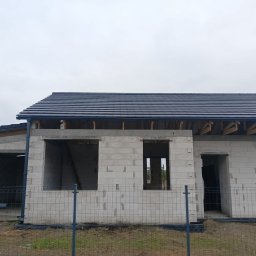 Usługi Ciesielsko-Dekarskie Daria Nawrocka - Surowy stan domu jednorodzinnego z betonu komórkowego, z częściowo ukończonym dachem z ciemnej dachówki ceramicznej, widoczne drewniane elementy konstrukcji dachu i orynnowanie, ogrodzenie...