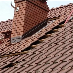 Usługi Ciesielsko-Dekarskie Daria Nawrocka - Częściowy demontaż pokrycia dachowego z dachówki ceramicznej wokół komina z cegły, widoczne łaty i kontrłaty, narzędzia dekarskie na dachu.