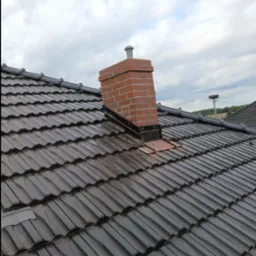 Ciemna dachówka ceramiczna z ceglanym kominem, zabezpieczonym obróbką blacharską, na tle pochmurnego nieba. W oddali widoczne gniazdo bocianie.