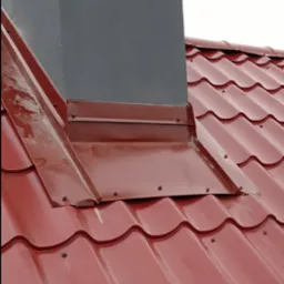 Czerwona dachówka ceramiczna z obróbką blacharską komina w kolorze bordowym, widoczne śruby mocujące.