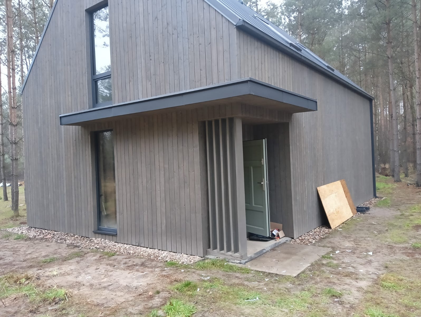 Nowoczesny dom z drewnianą elewacją w odcieniach szarości, z widocznym wejściem zadaszonym i pionowymi elementami dekoracyjnymi, otoczony lasem sosnowym.