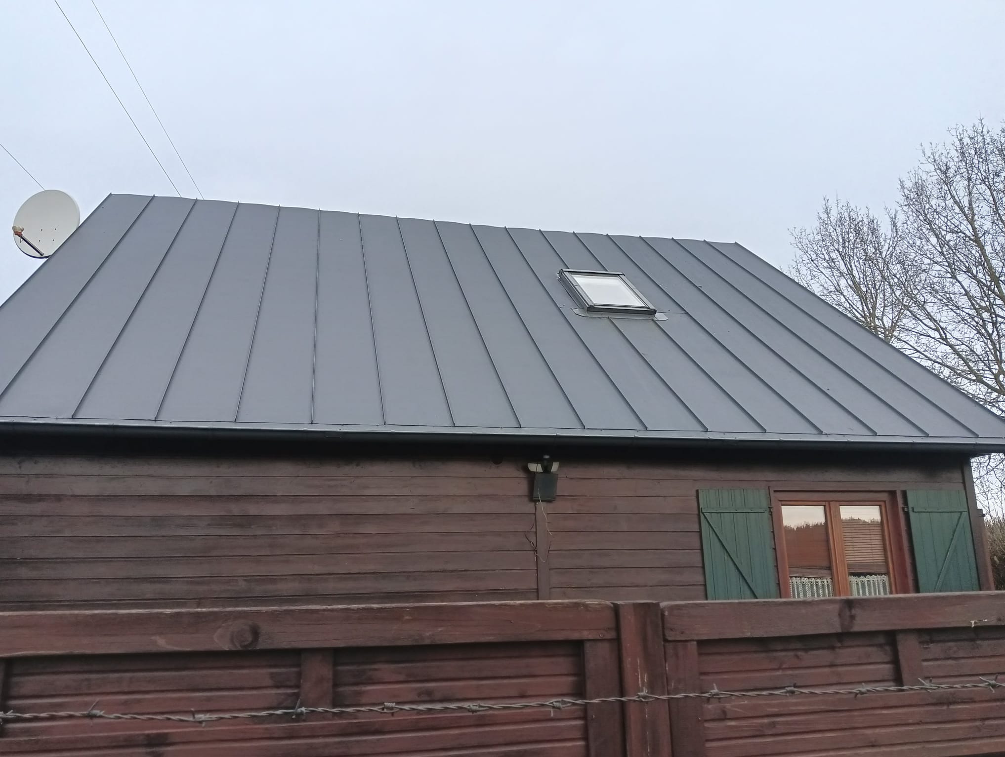 Szary dach z blachy trapezowej z oknem dachowym na drewnianym domu z brązowymi ścianami i zielonymi okiennicami, widoczna antena satelitarna.