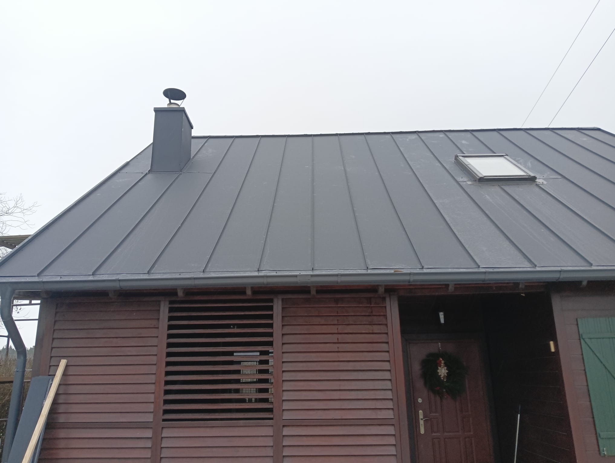 Dom z ciemnoszarym dachem z blachy na rąbek stojący, kominem i oknem dachowym, drewnianą elewacją i świątecznym wieńcem na drzwiach.