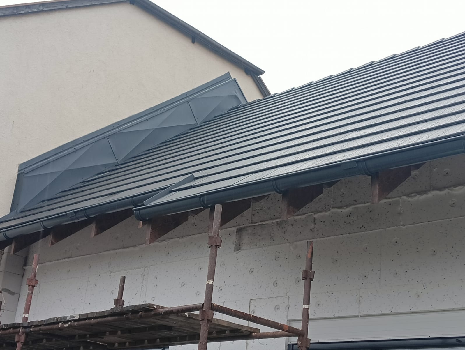Nowy dach z ciemnej dachówki ceramicznej, widoczne obróbki blacharskie na styku z sąsiednim budynkiem oraz rusztowanie przy elewacji z ociepleniem.
