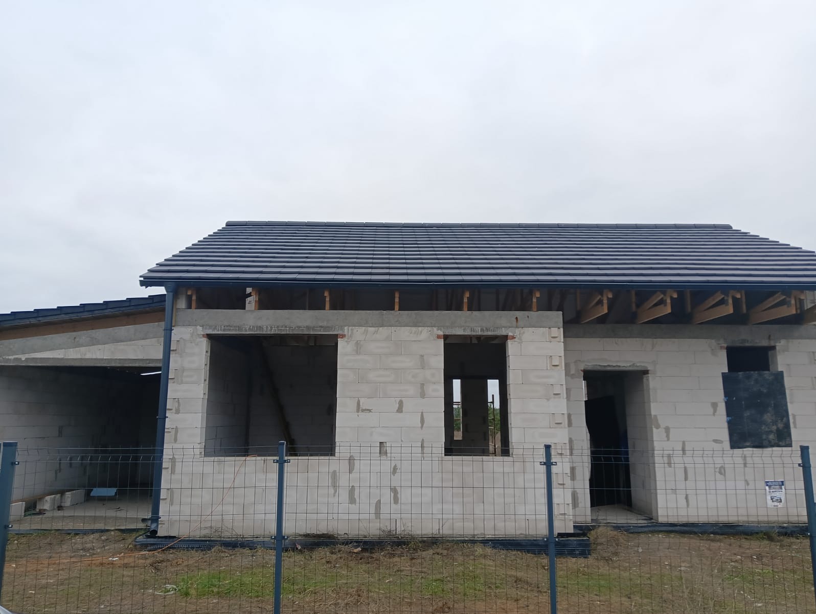 Surowy stan domu jednorodzinnego z betonu komórkowego, z częściowo ukończonym dachem z ciemnej dachówki ceramicznej, widoczne drewniane elementy konstrukcji dachu i orynnowanie, ogrodzenie...