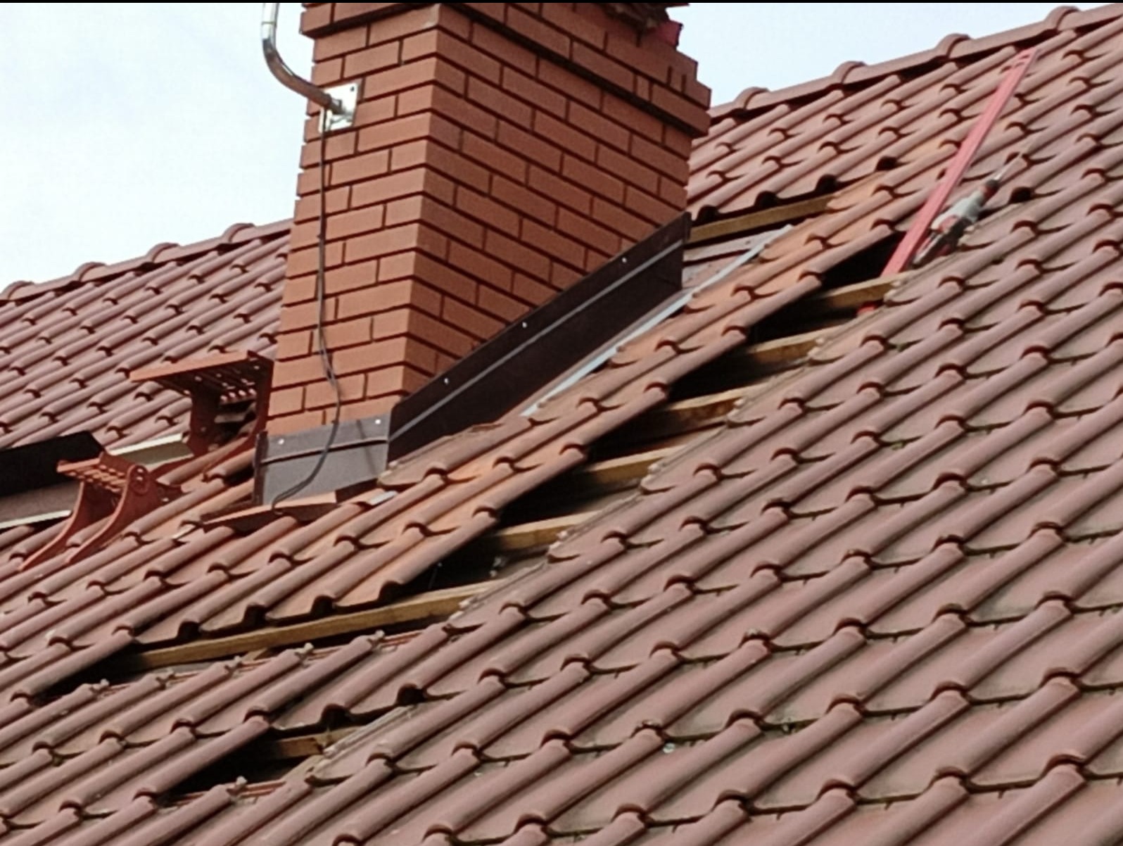 Częściowy demontaż pokrycia dachowego z dachówki ceramicznej wokół komina z cegły, widoczne łaty i kontrłaty, narzędzia dekarskie na dachu.