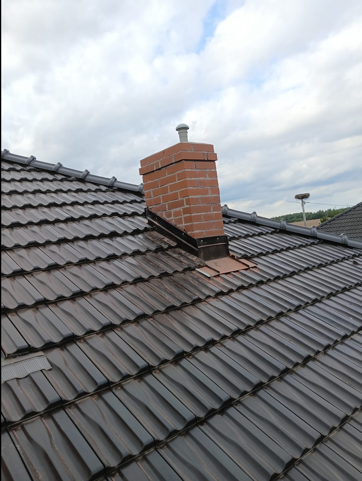 Ciemna dachówka ceramiczna na dachu domu z ceglanym kominem z metalowymi obróbkami, widok z góry na tle pochmurnego nieba.