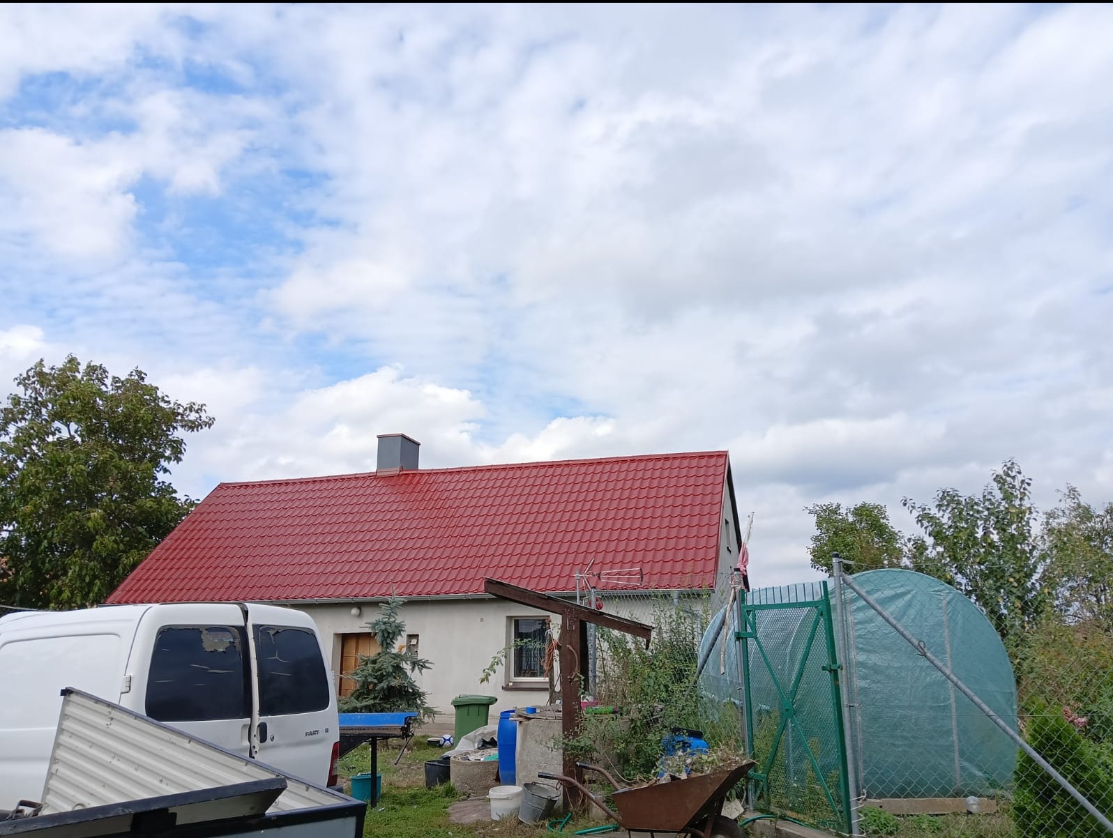 Dom jednorodzinny z nowym, czerwonym dachem z blachodachówki, widoczny z perspektywy podwórza z zaparkowanym białym busem i tunelem foliowym.