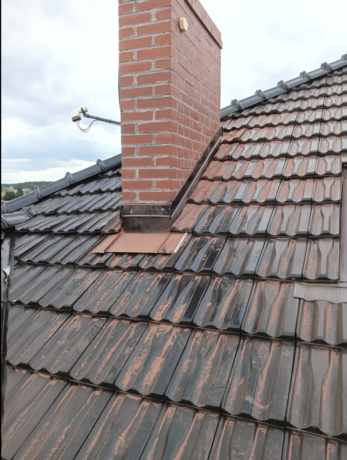 Ciemna dachówka ceramiczna z kominem z czerwonej cegły i anteną satelitarną, widok z góry na tle pochmurnego nieba.