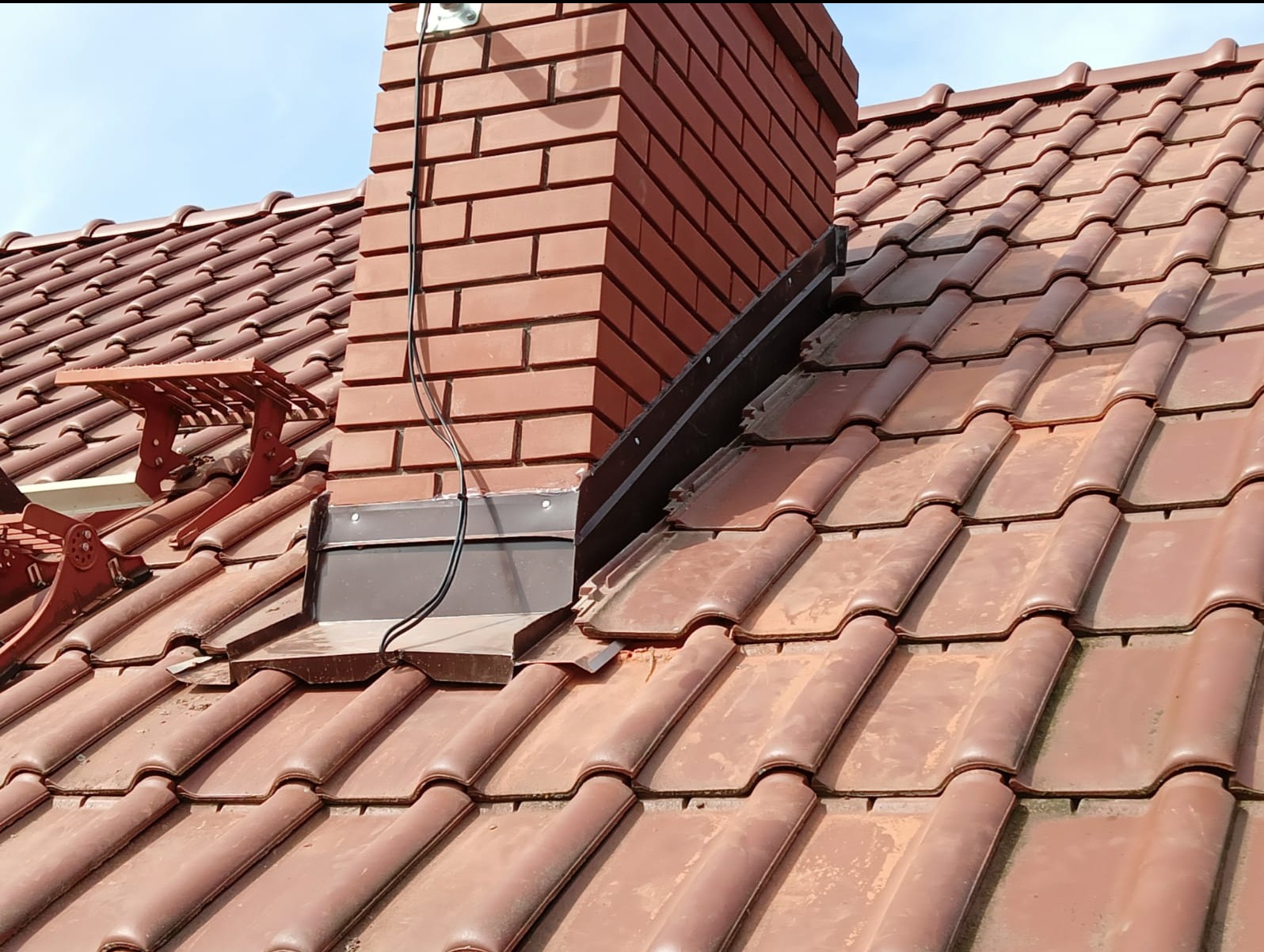 Ceglany komin wystający z dachu pokrytego czerwoną dachówką ceramiczną, widoczne obróbki blacharskie i uchwyt do montażu ławy kominiarskiej.