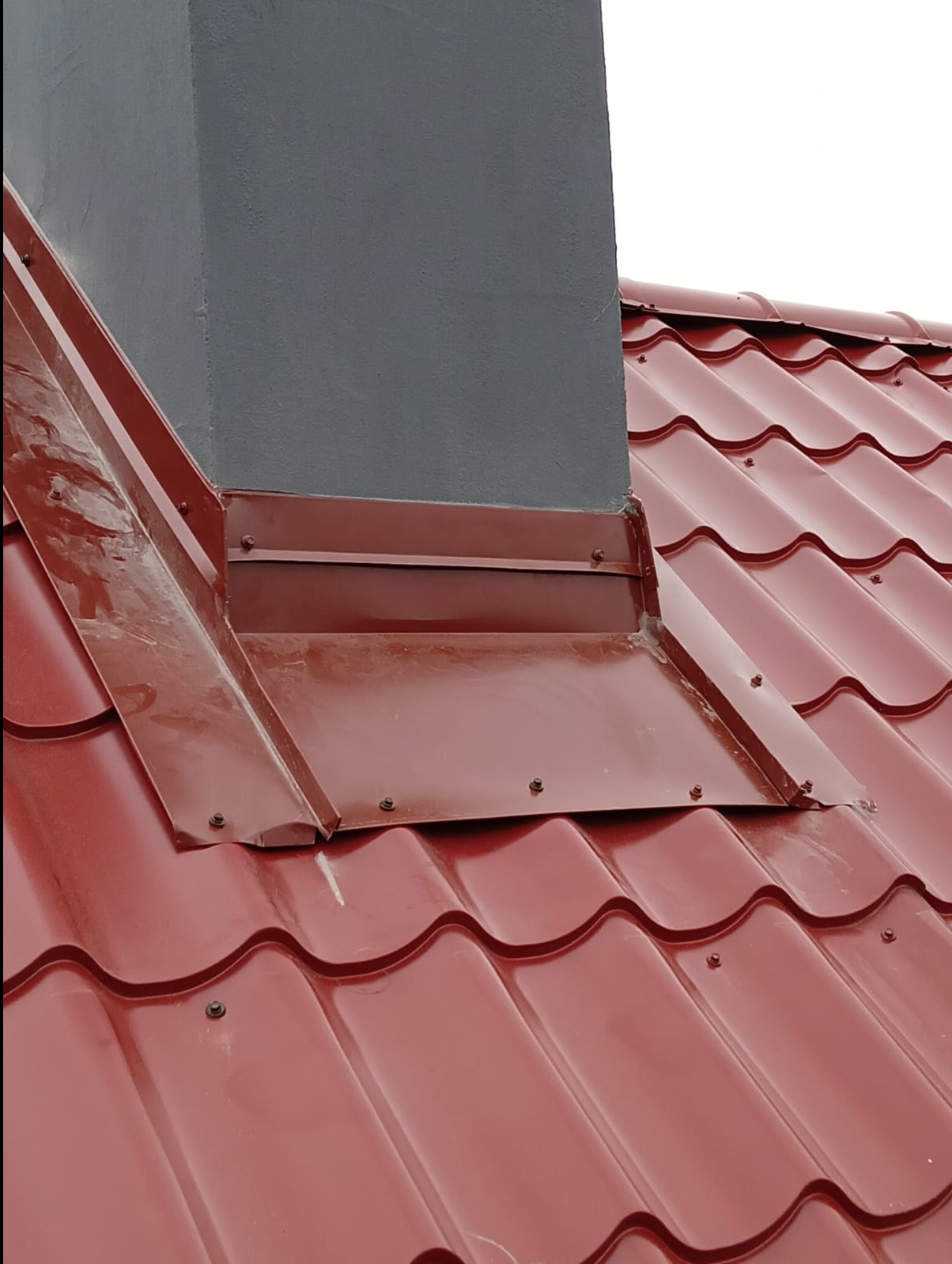 Czerwona dachówka ceramiczna z obróbką blacharską komina w kolorze bordowym, widoczne śruby mocujące.