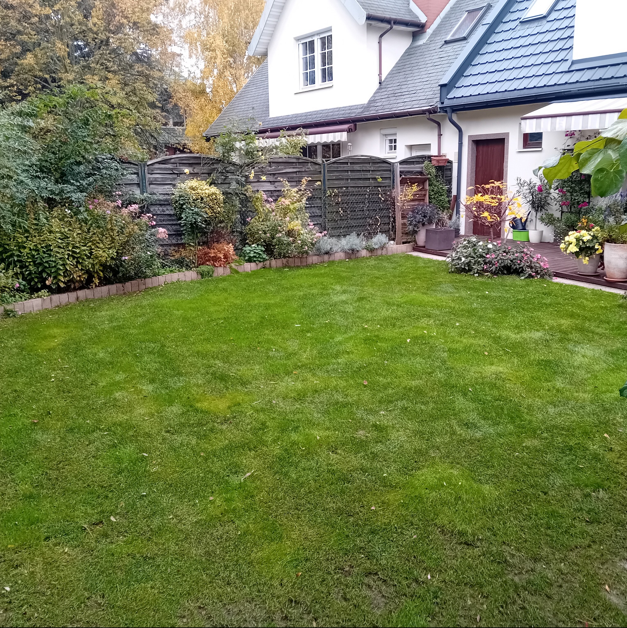 Widok na zadbany trawnik przed domem z drewnianym tarasem, rabatami kwiatowymi i ozdobnym ogrodzeniem.