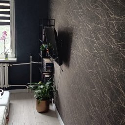 MARTINEZ CONSTRUCTION - Marcin Przytulski - Wnętrze pokoju po remoncie: ściana w ciemnobrązowej tapecie z jasnymi wzorami, jasna podłoga z paneli, czarna ściana obok okna i grzejnik.