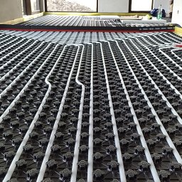 MARTINEZ CONSTRUCTION - Marcin Przytulski - Ułożona instalacja ogrzewania podłogowego w surowym wnętrzu z dużymi oknami. Białe rurki na czarnej macie, widok z perspektywy podłogi.