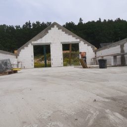 MARTINEZ CONSTRUCTION - Marcin Przytulski - Wznoszenie ścian budynku z białych bloczków, widoczne otwory okienne i drzwiowe. Prace murarskie w toku, widoczne rusztowania i narzędzia. Na drugim planie zieleń drzew.