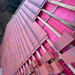 MARTINEZ CONSTRUCTION - Marcin Przytulski - Układanie dachówki karpiówki na różowej membranie, widoczne drewniane łaty i fragmenty dachówek w pakietach. Praca w toku, widok z dołu na dach.