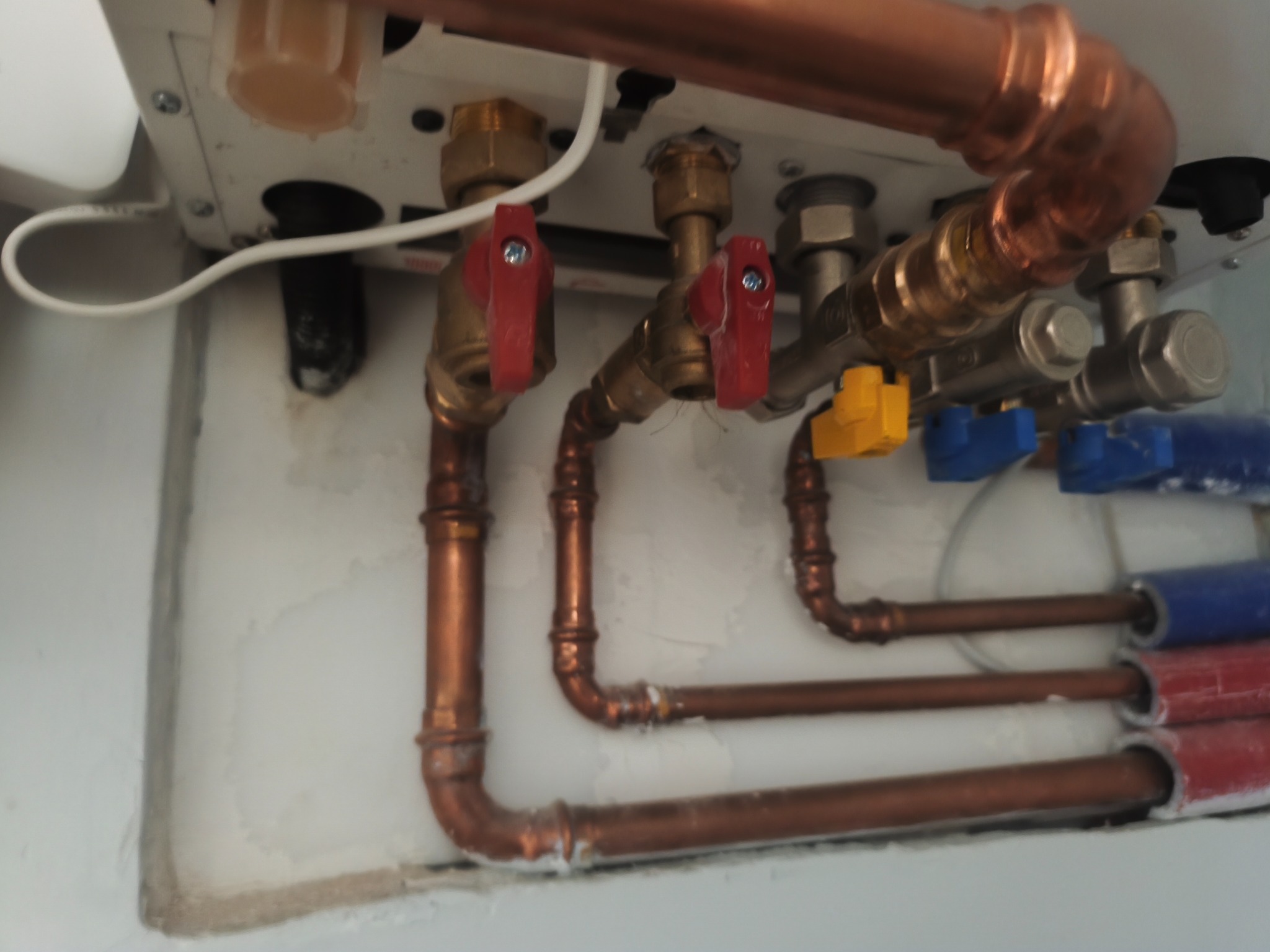 Miedziane rury instalacji wodnej z zaworami w kolorze czerwonym, żółtym i niebieskim, zamontowane na białej ścianie. Widoczne łączenia i fragment kotła.