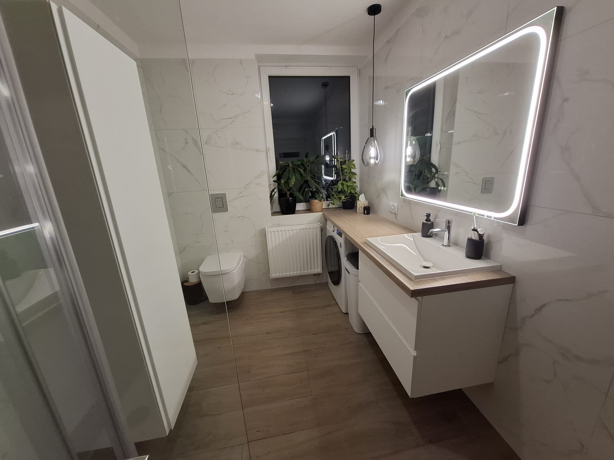 Nowoczesna łazienka z drewnianą podłogą i blatem, marmurowe płytki ścienne, podświetlane lustro i designerska lampa wisząca. Umywalka z szafką i pralka.