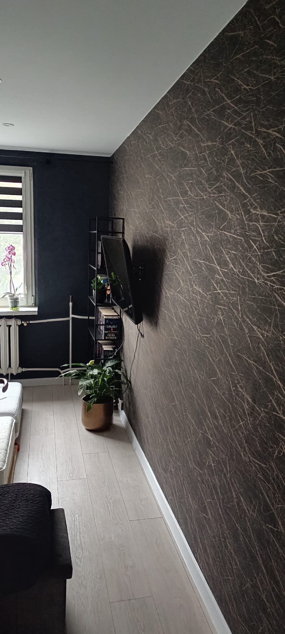 Wnętrze pokoju po remoncie: ściana w ciemnobrązowej tapecie z jasnymi wzorami, jasna podłoga z paneli, czarna ściana obok okna i grzejnik.